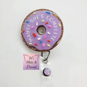 May include: Purple donut-shaped badge holder with gold glitter and white text that reads "Maybe She's Complete". The donut has pink, red, blue, and yellow sprinkles. The badge holder is attached to a clear retractable badge reel with a purple glitter charm.