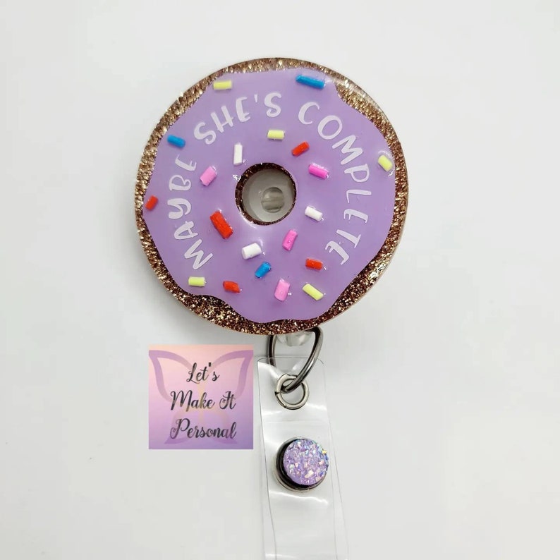 May include: Purple donut-shaped badge holder with gold glitter and white text that reads "Maybe She's Complete". The donut has pink, red, blue, and yellow sprinkles. The badge holder is attached to a clear retractable badge reel with a purple glitter charm.