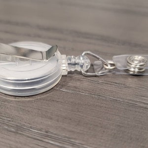 May include: A clear plastic retractable badge holder with a silver metal clip. The holder has a clear plastic strap with a silver metal button.
