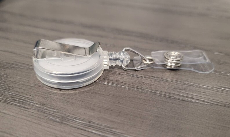 May include: A clear plastic retractable badge holder with a silver metal clip. The holder has a clear plastic strap with a silver metal button.