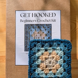 May include: A cardboard box containing a crochet kit. The box is labelled "GET HOOKED Beginners Crochet Kit". The kit includes a crochet hook, yarn, and instructions. The crochet square is blue, teal, and beige.