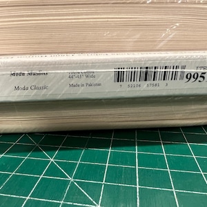 Puede incluir: Una pila de rollos de tela envueltos en plástico transparente. La etiqueta dice "Moda Muslins" y "Moda Classic". La tela mide 111.76cm-114.3cm de ancho y está hecha en Pakistán. También se ve un código de barras y el número "9951 12".