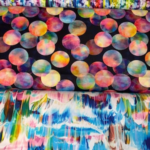 May include: Three stacked bolts of fabric. The top layer features a dense pattern of small white flowers on a vibrant, multi-colored vertical striped background. The middle layer displays colorful, watercolor-style circles on a black background. The bottom layer shows a fluid, marbled pattern of blue, pink, yellow, and white streaks.