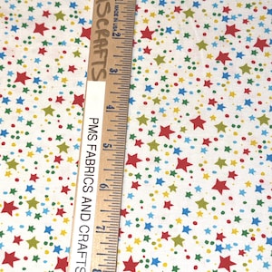 May include: A white fabric featuring a scattered pattern of red, blue, and lime green stars and small dots. A wooden ruler is placed vertically on the fabric, showing measurements in inches up to 8 inches, with the text PMSCRAFTS printed on it.