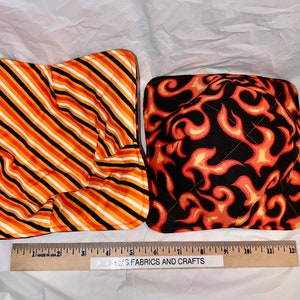 May include: Two quilted pot holders, one with orange and black stripes and the other with a flame pattern on a black background. The pot holders are on a white surface with a ruler that reads "Crafts Fabrics and Crafts".