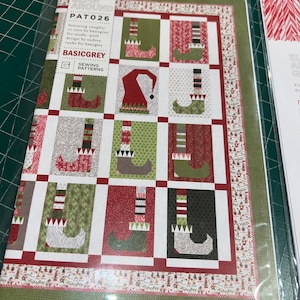 May include: A sewing pattern titled "Elfin Around" by Basicgrey. The pattern features a quilt design with elf boots and hats in red, green, and white. The pattern is in a clear plastic sleeve.
