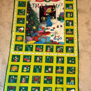 May include: A rectangular fabric panel featuring a Grinch-themed Christmas advent calendar. The design includes a central illustration of the Grinch by a fireplace with a tree and gifts, surrounded by numbered squares from 1 to 24 in green.