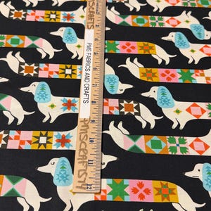May include: A black fabric featuring a repeating pattern of white dachshunds with light blue ears and colorful geometric quilt-style bodies. A wooden ruler measuring 12 inches is placed vertically over the fabric, labeled PMS FABRICS AND CRAFTS.