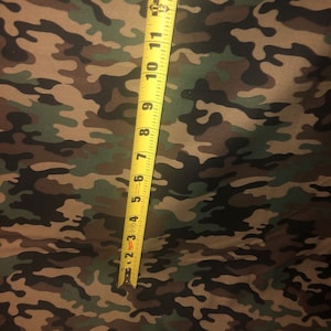 May include: A fabric featuring a camouflage pattern in shades of brown, black, and green. A yellow measuring tape is placed vertically over the fabric, showing a length measurement extending from zero to twelve inches on the tape's scale.