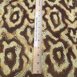 May include: Brown and yellow fabric with a repeating pattern of small circles. The fabric is textured and has a subtle sheen. A ruler is placed on the fabric, with the text "PMS FABRICS AND CRAFTS" visible.