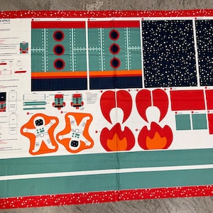 May include: A fabric panel with a space theme, featuring rocket ship components, astronauts, and flames in red, orange, teal, and navy blue. The panel includes the text "321 BLAST OFF BACKPACK" and is bordered with a red dotted pattern.