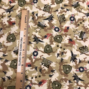 May include: Camouflage fabric with military-themed designs. The fabric features jets, stars, flags, and the text "USA Veteran Forces" and "Marine". A wooden ruler with the text "PMS Fabrics and Crafts" is placed on the fabric.