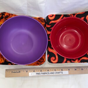 May include: Two bowls, one purple and one red, are sitting on top of two black and orange fabric pot holders. The pot holders have a flame design. A wooden ruler with the text "PMS Fabrics and Crafts" is in the bottom of the image.