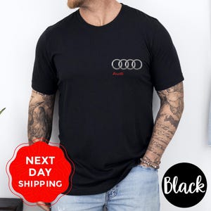 May include: A black short-sleeved t-shirt featuring the silver Audi four-ring logo on the left chest with the word Audi printed in red underneath. The shirt has a crew neckline and a standard fit, providing a clean and simple appearance.