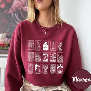 Może przedstawiać: Bordowa bluza z białymi ilustracjami kart tarota. Projekt zawiera r&oacute;żne karty tarota z tytułami takimi jak "The Eye", "The Dark" i "The End". Bluza jest w jednolitym bordowym kolorze.