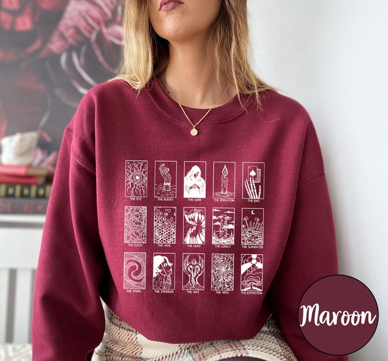 Może przedstawiać: Bordowa bluza z białymi ilustracjami kart tarota. Projekt zawiera r&oacute;żne karty tarota z tytułami takimi jak "The Eye", "The Dark" i "The End". Bluza jest w jednolitym bordowym kolorze.