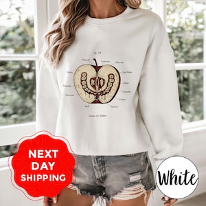 Könnte beinhalten: Weißes Sweatshirt mit einer detaillierten anatomischen Illustration eines Apfels mit Zähnen, beschriftet mit wissenschaftlichen Begriffen. Die Grafik ist in Braun- und Beigetönen gehalten. Das Sweatshirt hat einen Rundhalsausschnitt. Der Text "White" ist ebenfalls vorhanden.