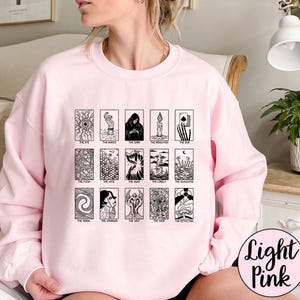Może przedstawiać: Jasnor&oacute;żowa bluza z nadrukiem kart tarota. Czarno-białe ilustracje kart tarota są ułożone w rzędach. Karty zawierają obrazy oka, postaci w płaszczu, świecy i innych. Słowa "Light Pink" są w okręgu po prawej stronie.