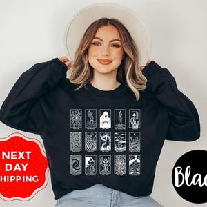 May include: Black sweatshirt featuring a white tarot card design. The design includes various tarot card illustrations with text. The sweatshirt is worn by a person wearing a cream-colored hat. The image also includes the text "NEXT DAY SHIPPING" and "Black".