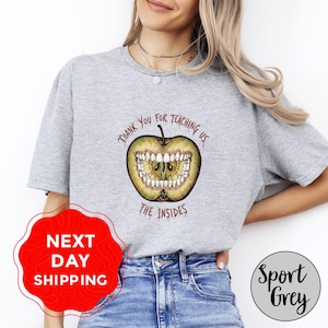 Könnte beinhalten: Hellgraues T-Shirt mit einem Apfel-Motiv mit Zähnen und dem Text "THANK YOU FOR TEACHING US THE INSIDES". Das Shirt trägt auch den Aufdruck "Sport Grey".