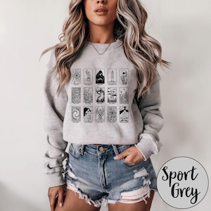 Może przedstawiać: Jasnoszara bluza z czarno-białym wzorem kart tarota. Bluza ma okrągły dekolt i długie rękawy. Słowa "Sport Grey" są wydrukowane na okrągłej etykiecie w prawym dolnym rogu.