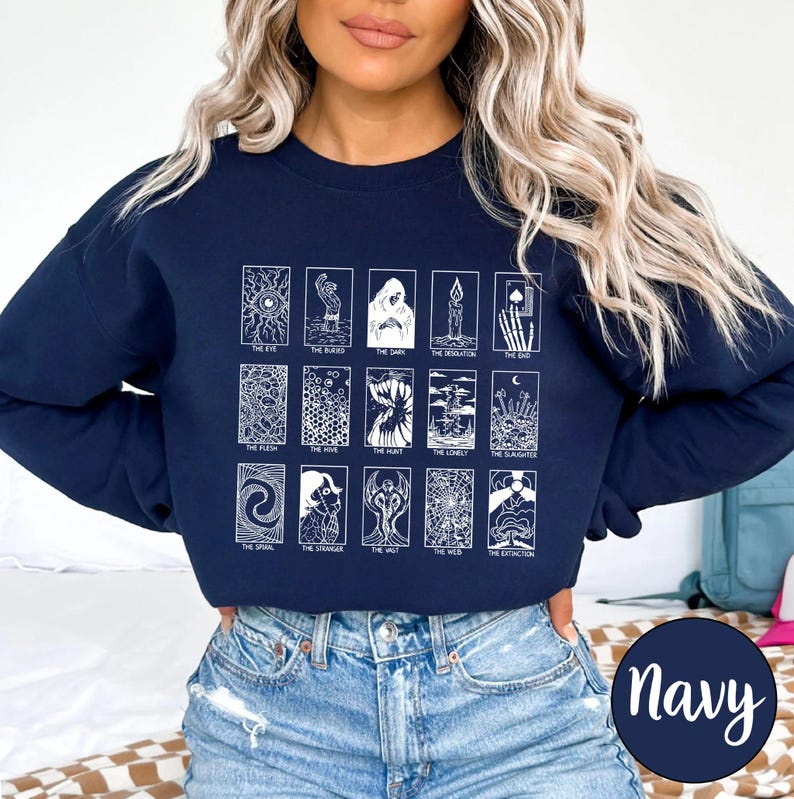 Może przedstawiać: Granatowa bluza z białymi ilustracjami kart tarota. Projekt zawiera r&oacute;żne obrazy kart tarota z tytułami takimi jak "The Eye", "The Bird" i "The End". Okrągła etykieta w prawym dolnym rogu głosi "Navy".
