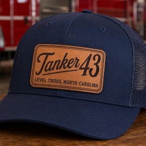May include: A navy blue trucker hat featuring a rectangular tan leather patch on the front. The patch is engraved with the text Tanker 43 and Level Cross, North Carolina in a dark brown font. The hat has a solid front panel and mesh back.