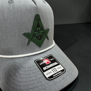 May include: Gray trucker hat with a white rope detail and a green and black emblem featuring a compass, square, and shamrock. The hat has a Richardson 112 Trucker tag.