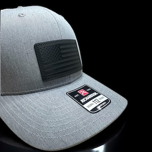 May include: A gray trucker hat with a black American flag patch. The hat features a curved brim and an adjustable strap. The Richardson brand label is visible on the front.