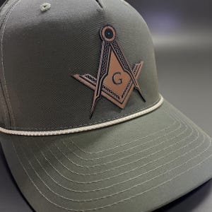 May include: Olive green baseball cap with a brown and black Masonic emblem. The emblem features a compass and square with the letter "G" in the center. The cap has a white rope detail along the brim.