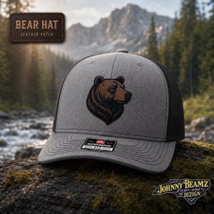 May include: A grey and black trucker hat featuring a brown bear head illustration on the front panel. The hat has a structured grey front, black mesh back, and a black brim with stitching. A sticker on the brim reads Richardson 112 Trucker.