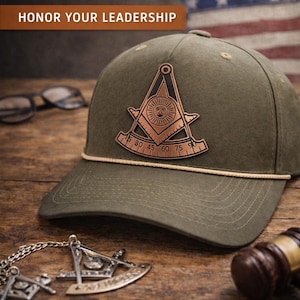 May include: An olive green snapback hat featuring a copper-colored patch of a square and compass with a sun face emblem. The hat includes a thin tan cord across the base of the crown and displays the text PAST MASTER FREEMASON HAT HONOR YOUR LEADERSHIP.
