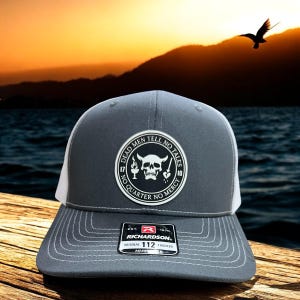May include: A gray and white trucker hat with a patch that reads "DEAD MEN TELL NO TALES" and "NO QUARTER NO MERCY". The hat has a curved brim and a mesh back. The hat is made by Richardson.