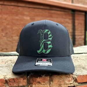 May include: Black trucker hat with a green and black American flag 'B' logo. The hat is adjustable and has a mesh back.