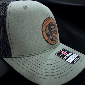 May include: A green and black trucker hat with a brown leather patch featuring a skull and crossbones with the text "Hatfield McCoy Devil Anse Trail" and a red, white, and blue Richardson logo with the text "112 Adjustable".