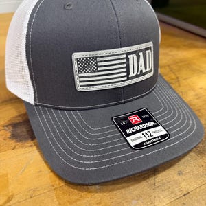 May include: A charcoal gray trucker hat with a white mesh back and a curved brim. The hat features a patch with an American flag design and the word "DAD". The Richardson brand tag is visible.