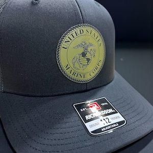 May include: A black trucker hat with a mesh back and a circular gold emblem featuring the United States Marine Corps logo. The hat has a curved brim and an adjustable strap. The Richardson tag is visible.