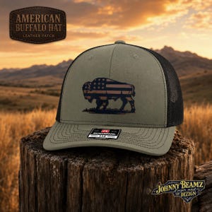 May include: An olive green trucker hat with a black mesh back and a structured front panel. The front features a dark graphic of a buffalo filled with a stars and stripes pattern. A small rectangular sticker is centered on the brim of the hat.