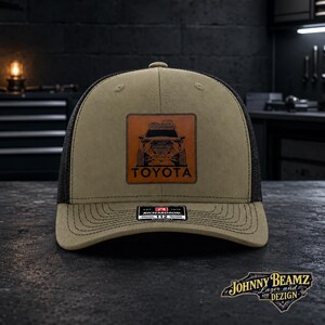 May include: A tan and black trucker hat featuring a square brown leather patch on the front. The patch displays a black graphic of a Toyota truck with the word TOYOTA printed below it. The hat has a structured crown and a curved brim with stitching.