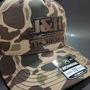 May include: A camouflage trucker hat with a brown and black logo that reads "J&H Welding and Fabrication LLC" and a phone number. The hat has a Richardson tag that says "Original 112 Trucker Adjustable".