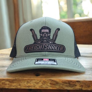 May include: A green trucker hat with a black mesh back. The front features a brown patch with the text "FREIGHTSHAKER" and a graphic of a skull with a handlebar mustache and exhaust pipes.