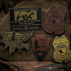May include: A collection of five tactical patches including a rectangular Jackson Hole grizzly bear sign, two shield-shaped Park Ranger badges, a National Park Service arrowhead patch, and a winged eagle emblem with a rifle, all on a brown base.