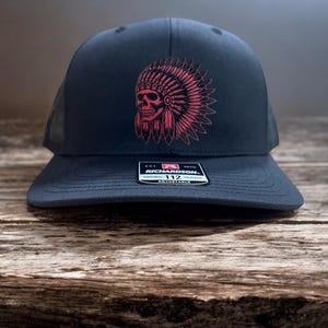 May include: A black trucker hat with a pink skull wearing a headdress. The hat has a mesh back and a black adjustable strap with a black label that reads "Richardson 112 Original Trucker Adjustable".
