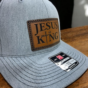 May include: A gray trucker hat with a brown leather patch that reads "JESUS is KING" in black lettering. The patch features a cross. The hat has a curved brim and an adjustable strap.