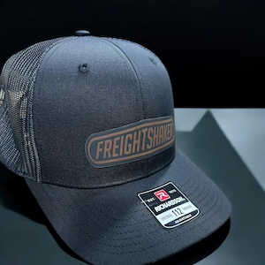 May include: Black trucker hat with a brown leather patch that says "FREIGHTSHAKER" in a white font. The hat has a black mesh back and a black adjustable strap with a white label that says "EST. 1910 RICHARDSON ORIGINAL 112 TRUCKER ADJUSTABLE".