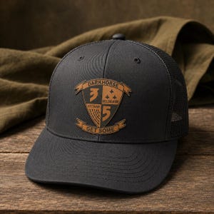 May include: A black baseball cap with a mesh back and a tan leather-like patch on the front. The patch features a shield design with the text DARKHORSE, 3, VIETNAM, BELLEAU WOOD, 5, and GET SOME, along with a small fleur-de-lis graphic.