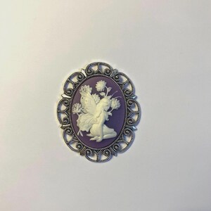 May include: Oval cameo brooch with a silver-tone filigree frame. The cameo features a white fairy with butterfly wings and flowers on a purple background. A decorative accessory.