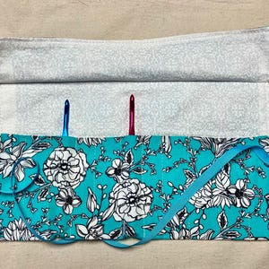 May include: A turquoise crochet hook roll with a floral pattern in white and black. The roll is secured with a light blue ribbon. Two crochet hooks, one blue and one pink, are visible inside the roll. The top flap is a light gray with a small pattern.