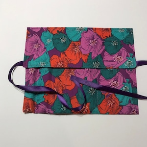 May include: A purple fabric pouch with a floral print in shades of pink, teal, and orange. The pouch has a purple ribbon tie closure.