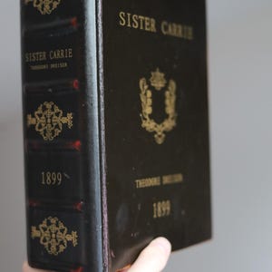 Puede incluir: Un libro antiguo de tapa dura titulado "Sister Carrie" de Theodore Dreiser, publicado en 1899. El libro tiene una cubierta oscura con letras doradas y detalles decorativos. La encuadernación muestra signos de desgaste.